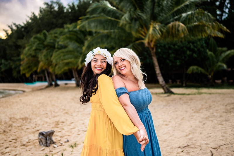 Family Photographer, two girls, friends and sisters, are back-to-back holding hands on the beach